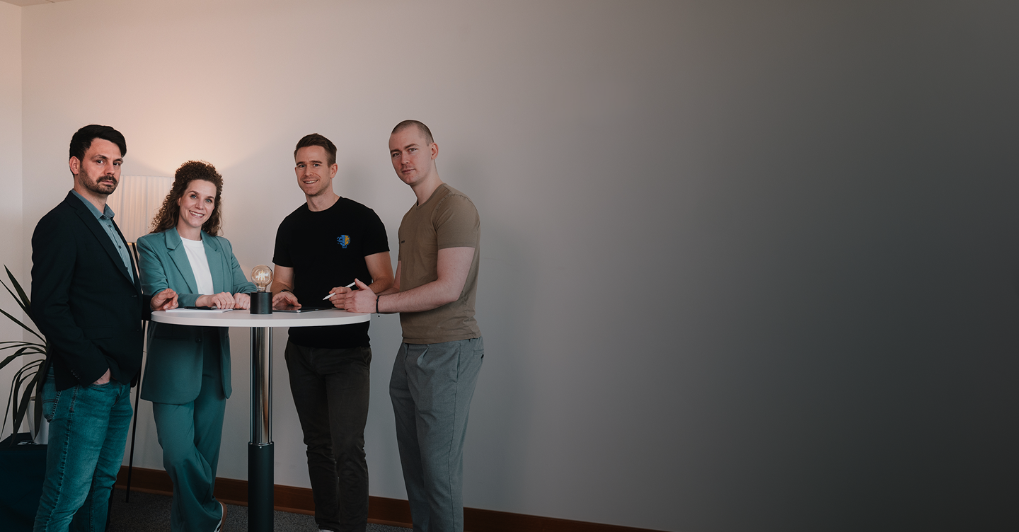 Von Links: Florian Bobek, Lorena Tippelt, Dr. Florian Imgrund und Konstantin Auffinger. Sie stehen um einen Stehtisch auf dem sich eine Lampe befindet. Links neben den Personen steht eine Pflanze.