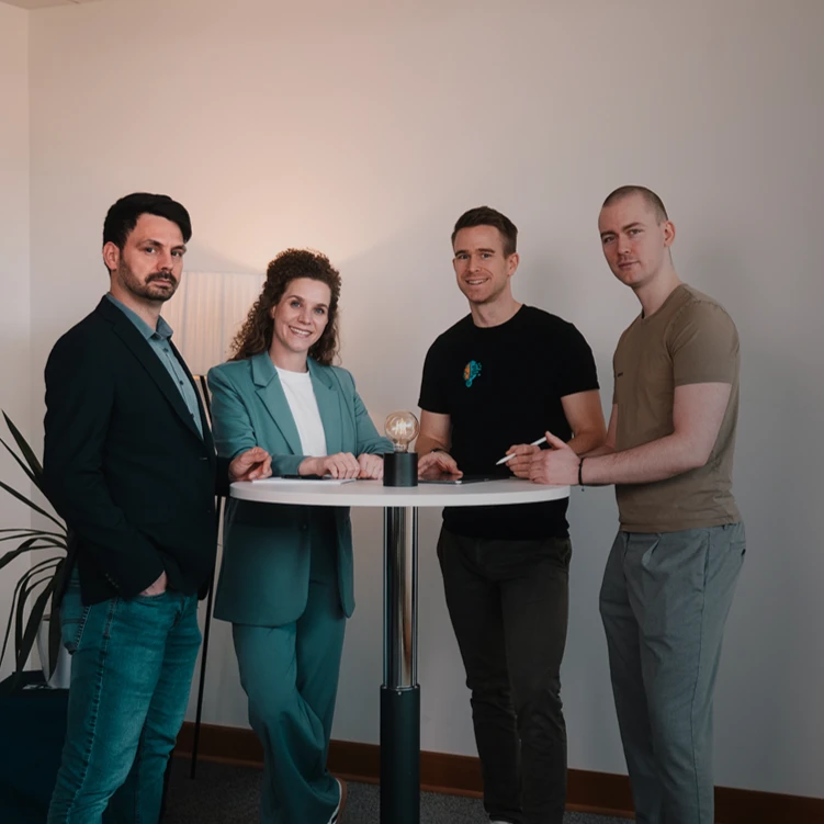 Von Links: Florian Bobek, Lorena Tippelt, Dr. Florian Imgrund und Konstantin Auffinger. Sie stehen um einen Stehtisch auf dem sich eine Lampe befindet. Links neben den Personen steht eine Pflanze.