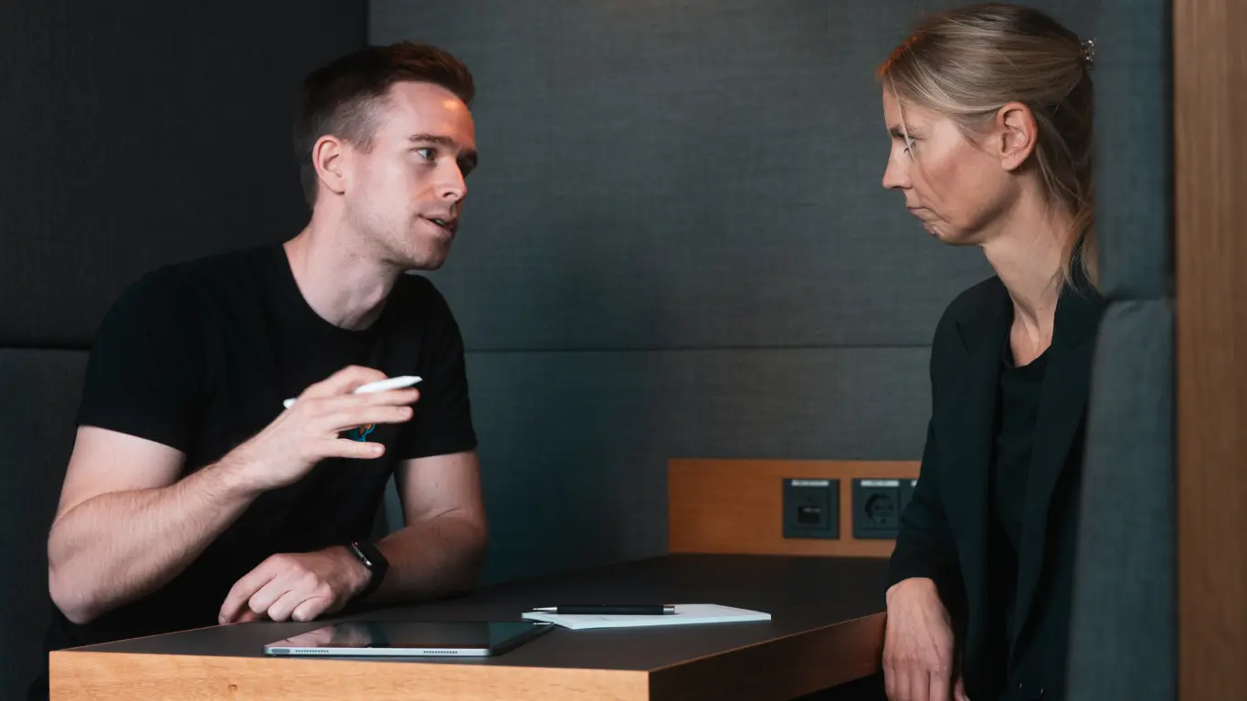 Dr. Florian Imgrund mit einem Kunden im Gespräch. Beide sitzen an einem Tisch, auf diesem ist ein Tablet, ein Notizblock und ein Stift zu sehen.