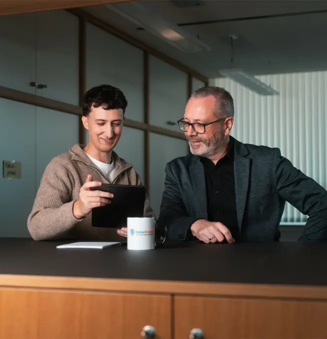 Tobias Reuss mit einem Kunden. Beide stehen an einem Stehtisch auf dem eine Tasse mit dem InnoBrain Logo, ein Stift und ien Block zu sehen sind. Tobias hält ein Tablet in den Händen und beide schauen hinein.