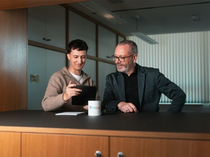 Tobias Reuss mit einem Kunden. Beide stehen an einem Stehtisch auf dem eine Tasse mit dem InnoBrain Logo, ein Stift und ien Block zu sehen sind. Tobias hält ein Tablet in den Händen und beide schauen hinein.