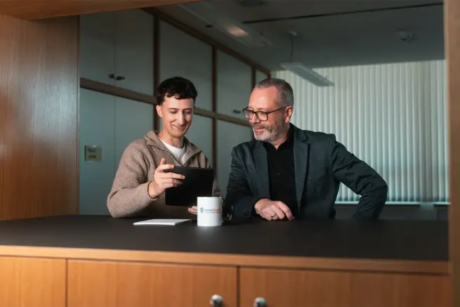 Tobias Reuss mit einem Kunden. Beide stehen an einem Stehtisch auf dem eine Tasse mit dem InnoBrain Logo, ein Stift und ien Block zu sehen sind. Tobias hält ein Tablet in den Händen und beide schauen hinein.