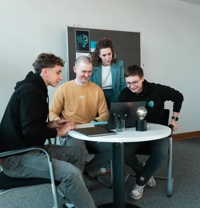 Lorena Tippelt, Konstantin Auffinger, Daniel Goncharov und Bruno Görß. Bruno, Konstantin und Daniel sitzen. Lorena steht hinter ihnen. Hinter Lorena steht ein Pinboard. Alle schauen in einen aufgeklappten Laptop. Der Laptop steht auf einem Tisch, worauf der Laptop, eine Lampe, ein Stiftehalter mit Stiften und ein weiterer Laptop vorzufinden sind.