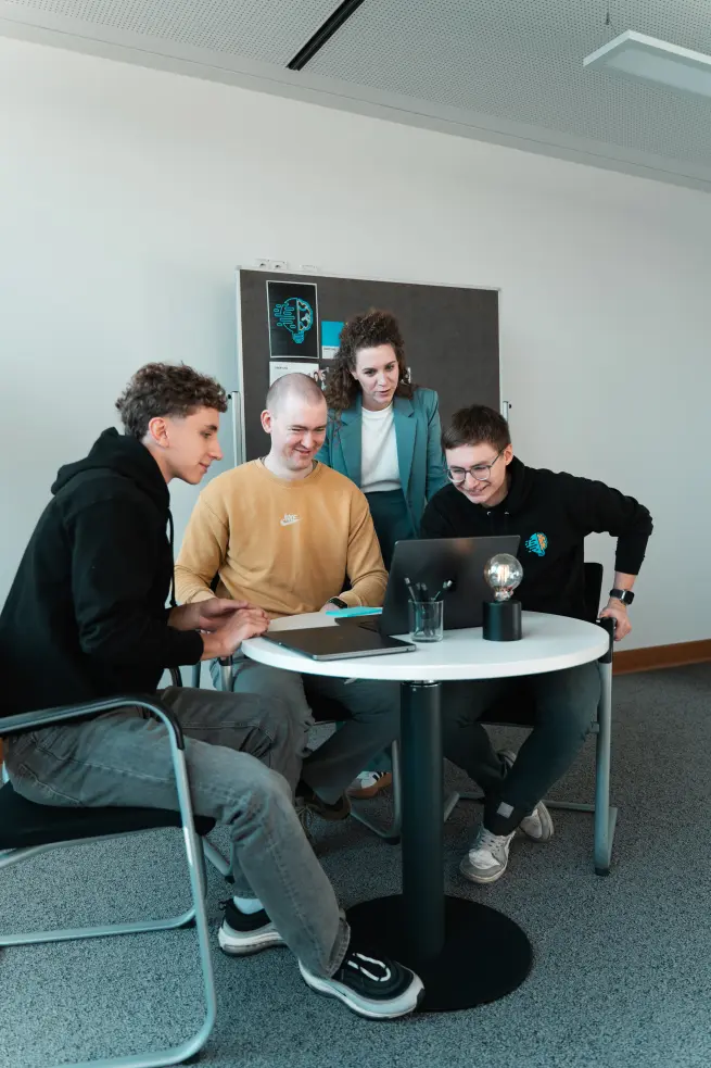Lorena Tippelt, Konstantin Auffinger, Daniel Goncharov und Bruno Görß. Bruno, Konstantin und Daniel sitzen. Lorena steht hinter ihnen. Hinter Lorena steht ein Pinboard. Alle schauen in einen aufgeklappten Laptop. Der Laptop steht auf einem Tisch, worauf der Laptop, eine Lampe, ein Stiftehalter mit Stiften und ein weiterer Laptop vorzufinden sind.