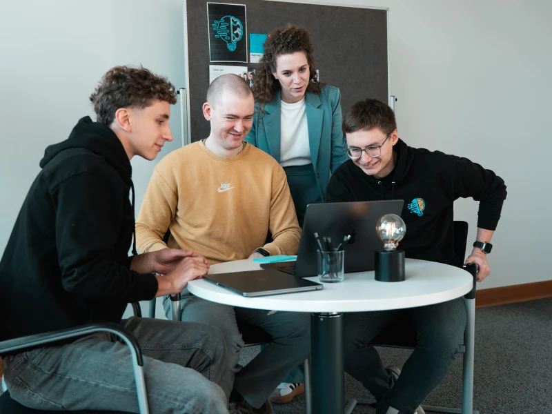 Lorena Tippelt, Konstantin Auffinger, Daniel Goncharov und Bruno Görß. Bruno, Konstantin und Daniel sitzen. Lorena steht hinter ihnen. Hinter Lorena steht ein Pinboard. Alle schauen in einen aufgeklappten Laptop. Der Laptop steht auf einem Tisch, worauf der Laptop, eine Lampe, ein Stiftehalter mit Stiften und ein weiterer Laptop vorzufinden sind.