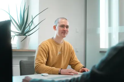 Konstantin Auffinger sitzt an einem Tisch mit ienem Kunden. Vor ihm sind Maus und Tastatur. Seine Hände sind an der Tastatur, sein Blick jedoch beim Kunden.