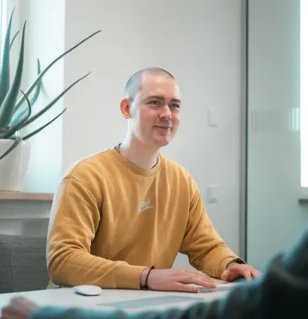 Konstantin Auffinger sitzt an einem Tisch mit ienem Kunden. Vor ihm sind Maus und Tastatur. Seine Hände sind an der Tastatur, sein Blick jedoch beim Kunden.