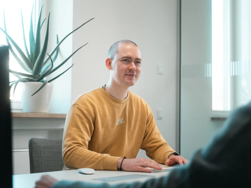 Konstantin Auffinger sitzt an einem Tisch mit ienem Kunden. Vor ihm sind Maus und Tastatur. Seine Hände sind an der Tastatur, sein Blick jedoch beim Kunden.