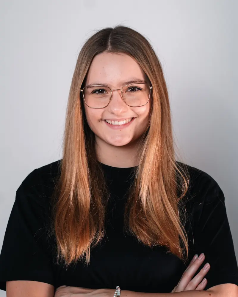 Emma Martin in einem Portraitformat. Sie trägt eine Brille, verschränkt ihre Arme und hat zwei Perlenketten am Handgelenk.